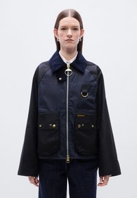 Ναυτικό μπουφάν με γιακά από κορδόνι, κλείσιμο με φερμουάρ, δύο μπροστινές τσέπες, χρυσές λεπτομέρειες και μαύρα μανίκια. Το υλικό φαίνεται ανθεκτικό και ανθεκτικό στις καιρικές συνθήκες.