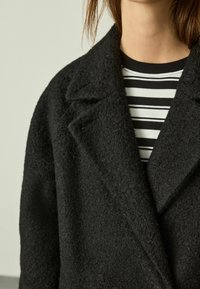 Person wearing a textured black overcoat over a black and white striped shirt, cropped at the neck and shoulders.