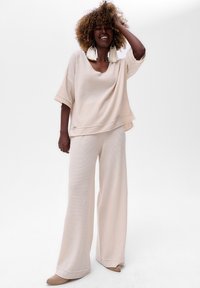 Vrouw in beige gebreide wijd uitlopende broek en een losse top met korte mouwen, glimlachend met haar hand in kroezig haar, met grote kwastoorbellen.