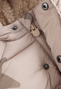 Doudoune beige avec une doublure en polaire douce et texturée. Dispose d'une tirette de fermeture en forme d'ours et de boutons-pression argentés.