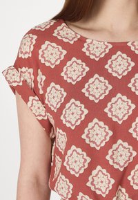 Haut à manches courtes en tissu brun rougeâtre avec un motif floral en crème et rose, doté d'un col rond et de manches retroussées.