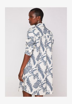 Donna con i capelli corti che indossa un abito bianco con motivi di pavoni blu e floreali, con un braccio leggermente piegato contro uno sfondo neutro.