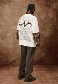 Prevelika bijela majica s grafičkim tekstom na leđima, složena preko smeškastog kariranog košulje, u kombinaciji s tamnosivim širokim hlačama i crnim cipelama.