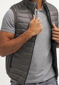 Harmaa toppaliivi, jossa on korkea kaulus, vetoketjukiinnitys ja sivutaskut, puettuna harmaan poolopaidan päälle. Sileä pinta, tikattu kuvio.