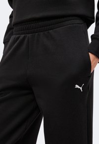 Κοντινή λήψη μαύρων παντελονιών Puma με ελαστική ζώνη και χέρι στην δεξιά τσέπη που δείχνει το λευκό λογότυπο Puma στον μηρό.