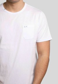 T-shirt bianco a maniche corte con una piccola tasca sul lato sinistro del petto. Presenta cuciture blu minimaliste sulla tasca. Tessuto in cotone morbido.