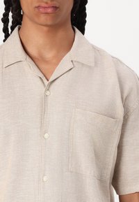 Chemise à manches courtes, beige, en tissu texturé, avec une poche poitrine et un col doux. Détails de couture subtils visibles.