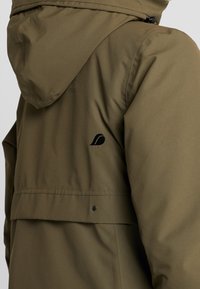 Veste à capuche vert olive en tissu texturé, avec un logo noir sur l'épaule et des détails à pression près de l'ourlet.