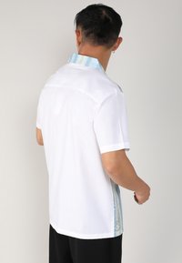 Chemise blanche à manches courtes avec un col texturé bleu clair et vert et des accents sur les côtés. Conçue en tissu léger.