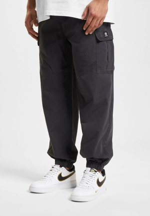 Black cargo pants with a relaxed fit, elastic cuffs, and side pockets. Worn with white sneakers featuring a black and gold swoosh.