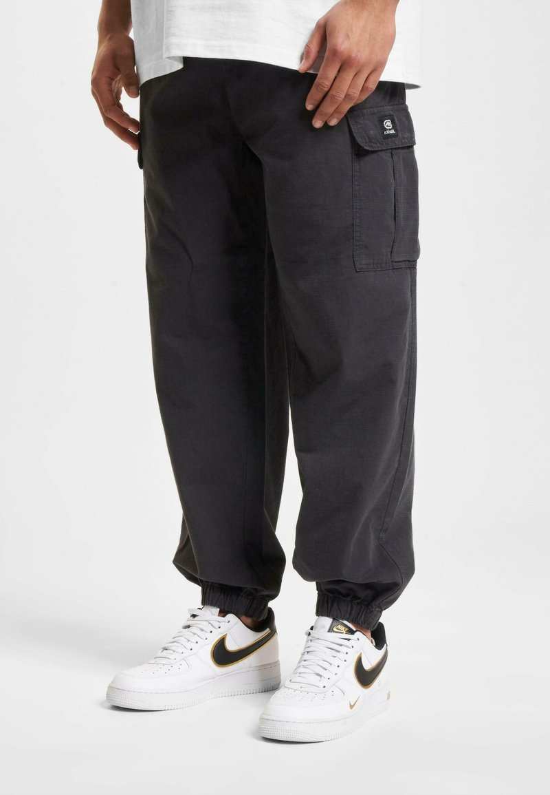 Zwarte cargobroek met een ontspannen pasvorm, elastische boorden en zijzakken. Gemaakt in combinatie met witte sneakers met een zwarte en gouden swoosh.