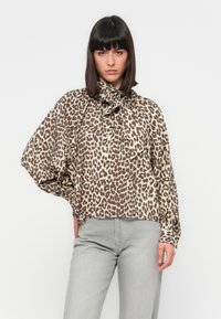 Blusa con stampa leopardata in beige e marrone, con colletto alto a fiocco, maniche lunghe a sbuffo e chiusura frontale con bottoni. Pantaloni grigi.