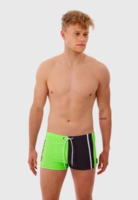 Un short de bain sportif présente un design à fentes en néon vert et noir, avec des rayures blanches et une fermeture à cordon à la taille.
