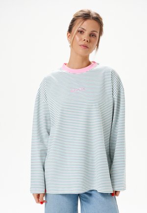 Femme aux cheveux attachés portant une chemise à manches longues rayée bleu clair et blanc avec un col rose et un jean bleu clair, debout devant un fond blanc.