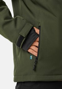 Grüne Softshelljacke mit einer Reißverschlusstasche, fleecegefütterten Bündchen und einem kleinen schwedischen Flaggenakzent. Der Stoff zeigt eine glatte, strukturierte Oberfläche.