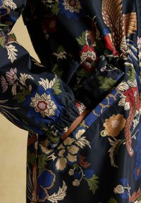 Mână purtând o jachetă bleumarin cu imprimeu floral, manșetă elastică, mână introdusă într-un buzunar frontal mare cu închidere cu nasture capsa și imprimeu botanic colorat.