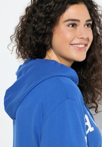 Felpa con cappuccio blu con coulisse, realizzata in tessuto morbido. Logo visibile sul davanti, con una vestibilità comoda e capelli ricci e strutturati sullo sfondo.