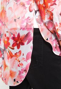 Blusa trasparente floreale in tonalità rosa e arancione, caratterizzata da un materiale leggero, un design fluido e un orlo curvo, abbinata a jeans scuri.