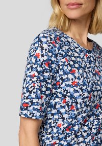 Kurzarm-Oberteil in blau, weiß und rot mit floralem Muster, mit rundem Ausschnitt und strukturiertem Stoff. Teilansicht einer Schulter.