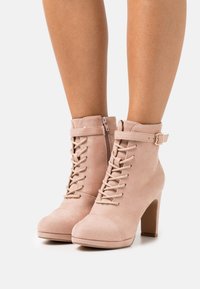 Růžové ankle boots z nubuku s kulatou špičkou, šněrováním vpředu, bočním zipem a ozdobným páskem s přezkou na kotníku. Podpatek o výšce 7,5 cm.