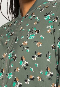 Blouson aviateur fleuri en tissu vert olive. Présente de petites fleurs multicolores en blanc, noir, sarcelle et marron. Fermeture éclair à l'avant, col côtelé.