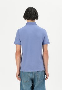 Polo tričko v světle modré barvě z bavlny, krátké rukávy a standardní límec. Zadní pohled ukazuje jednoduchý design bez viditelných log nebo vzorů.