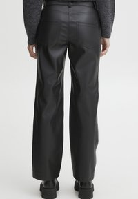 Pantaloni a gamba larga in pelle nera con una superficie liscia, dotati di quattro tasche e dettagli di cucitura visibili. Abbinati a calzature nere.