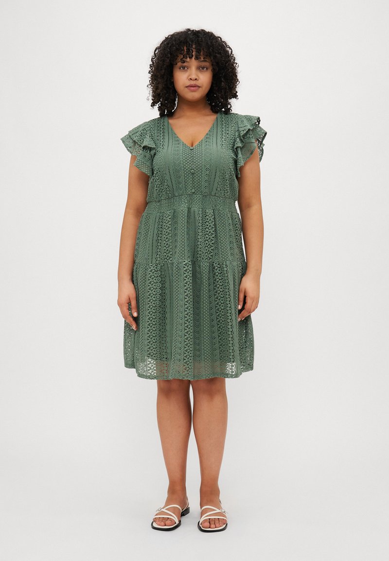 Green lace dress with short ruffled sleeves, a V-neckline, and tiered skirt. Worn with white sandals, featuring delicate straps.