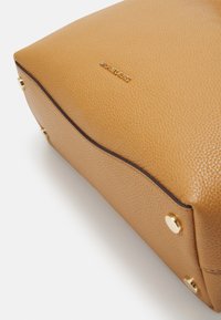 Kč medová kožená kabelka s texturovaným povrchem, zdůrazněná zlatými kovovými prvky a logem vyraženým na boku. Kulatý tvar, jemné hrany.