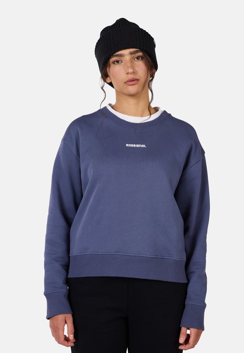 Niebieska bawełniana bluza z luźnym krojem, z prążkowanymi mankietami i dołem. Posiada białe logo na piersi. Noszona z czarną dzianinową czapką.