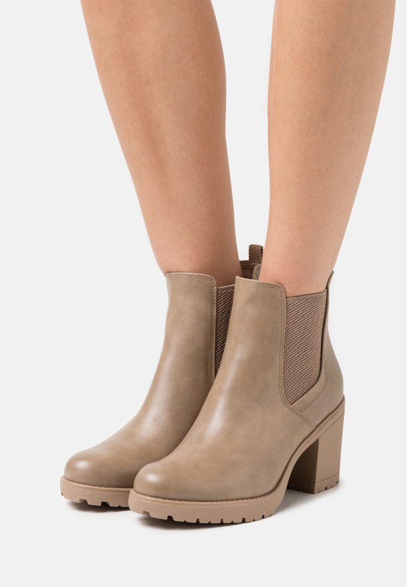 Bottines en cuir marron clair à talons épais portées sur des jambes nues sur un fond blanc uni.