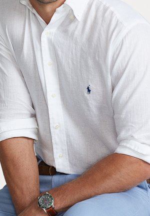 Chemise blanche en lin à manches retroussées, arborant un petit logo bleu sur la poitrine, associée à un pantalon bleu clair et à une montre verte.
