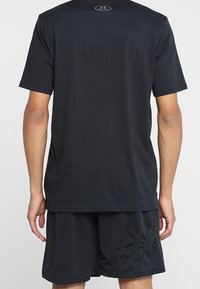 Hombre con una camiseta de manga corta negra lisa y pantalones cortos deportivos de malla negros, visto desde atrás sobre un fondo blanco.