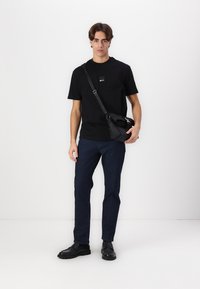 Svart crew neck t-skjorte med en liten logo, blå jeans og svarte lær-sko. Modellen holder en svart messenger-bag.
