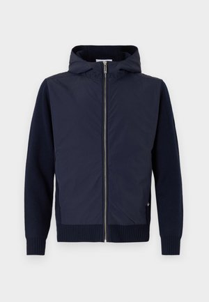 La chaqueta con capucha en color navy combina una capa exterior suave y ligera con mangas y dobladillos de punto acanalado. Cierre frontal con cremallera y un sutil detalle de logo.