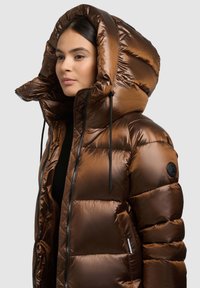 Brūna pūkaina jaka ar spīdīgu apdari, lielu kapuci, vertikālu šūšanu, melnām auklām un logotipa plāksteri uz piedurknes.