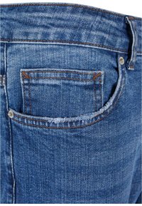 Vaqueros de mezclilla con un lavado azul oscuro, que presentan un bolsillo trasero, detalles de costura en naranja y una textura ligeramente desgastada.