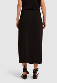 Schwarzer, knöchellanger Rock mit geradem Design und glatter Textur. Verfügt über minimale Nähte. Kombiniert mit glänzenden schwarzen Schuhen, die mit Schleifen verziert sind.