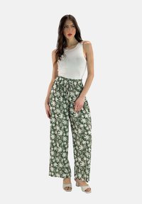 Groene bloemenbroek met een losse pasvorm, met een tailleband met trekkoord en een patroon van witte madeliefjes. Gepaard met een mouwloze witte top en lichte schoenen.