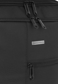 Czarna torba z teksturowanej tkaniny z podwójnymi czarnymi suwakami, przednią kieszenią i srebrną metką z logo. Skupia się na funkcjonalności i minimalistycznym designie.