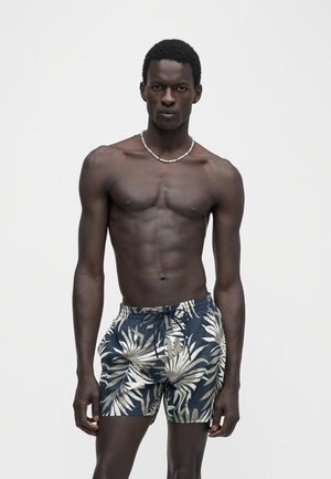 Homme à la peau foncée avec des cheveux courts et bouclés, portant un short de bain bleu et beige à imprimé tropical et un collier de perles, debout devant un fond uni.