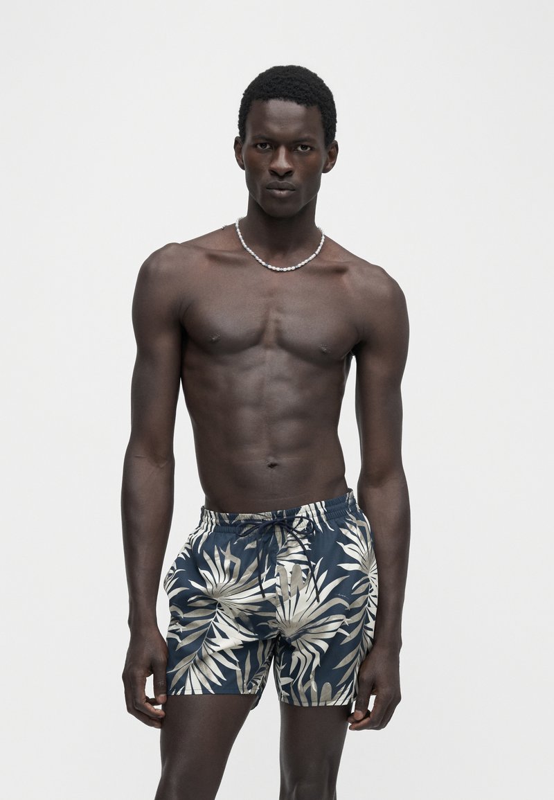 Homme à la peau foncée avec des cheveux courts et bouclés, portant un short de bain bleu et beige à imprimé tropical et un collier de perles, debout devant un fond uni.