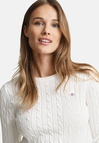 Pull en maille blanc avec un motif torsadé, doté d'un col rond et d'un petit logo sur la poitrine. Texture douce et coupe ajustée.