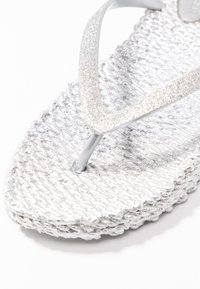 Silver flip-flop med en texturerad, vävd sula och ett glittrigt, triangulärt band. Designen är minimalistisk med en slät, blank yta.