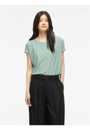 Femme aux longs cheveux foncés portant un haut court à manches courtes vert clair en eyelet, rentré dans un pantalon noir taille haute à jambes larges, debout sur un fond blanc.