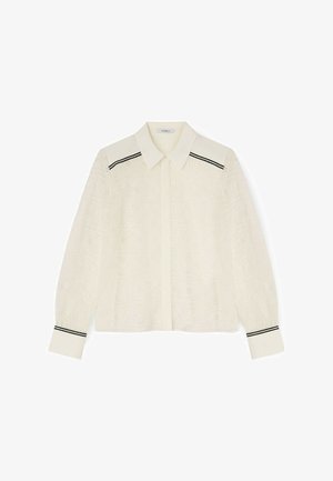 Blouse en dentelle crème à manches longues avec un col pointu, une patte de boutonnage, et des doubles bandes noires sur les épaules et les poignets.