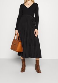 Sort ribbet langærmet kjole med plisseret nederdel og bindebånd i taljen. Brun struktureret håndtaske og teksturerede ankelstøvler.