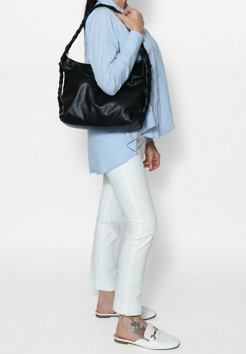 Borsa a spalla in pelle nera con manici intrecciati, caratterizzata da una texture liscia e un design spazioso, tenuta da una persona vestita con una camicia blu e pantaloni bianchi.