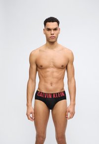 Άνδρας χωρίς πουκάμισο με κοντά σγουρά μαλλιά, στέκεται, φοράει μαύρα εσώρουχα Calvin Klein με κόκκινο λογότυπο στη μέση, μπροστά σε απλό φόντο.