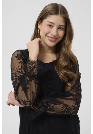 Young woman with long wavy hair smiles, wearing a black lace top with sheer floral sleeves and gold hoop earrings.